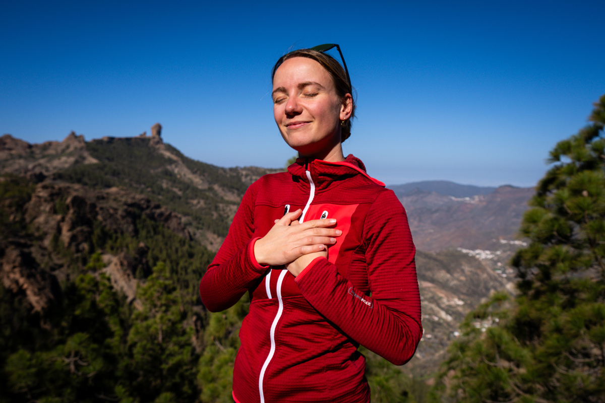 Angie, la facilitadora, con los ojos cerrados y las manos en el pecho, en una postura de meditación o canalización energética, con el Roque Nublo de Gran Canaria al fondo. La imagen refleja una profunda conexión espiritual con la naturaleza.